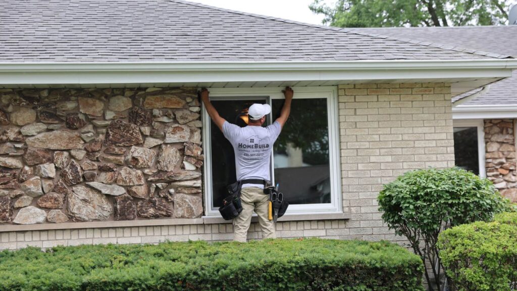 A person installs a window on a house with stone and brick exterior.